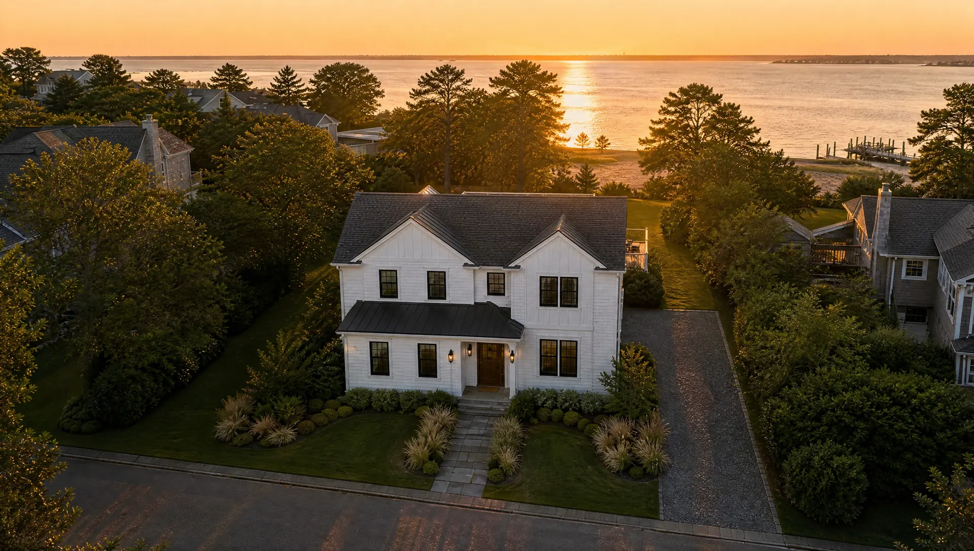 Aerial view of a white coastal home with a dark roof at sunset, the bay beyond the treeline.